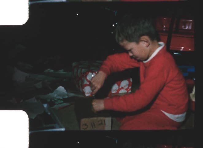 A young boy wearing bright red pajamas is kneeling on the floor unwrapping a gift with red and white patterned paper he smiles widely at the camera after successfully unwrapping the present which is surrounded by scattered wrapping paper and ribbons