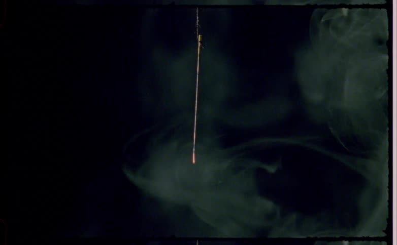 A thin stick like object hangs vertically as smoke rises against a dark background