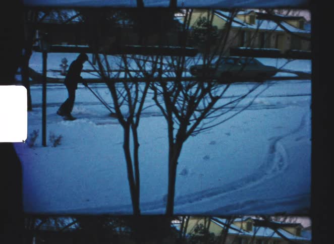 A man in silhouette shovels snow off a residential sidewalk during a winter day surrounded by bare trees and snowcovered ground