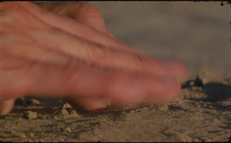 A very closeup shot focuses on a persons hand touching dry dirt eventually using their fingertips to pick up a thin red string from the ground