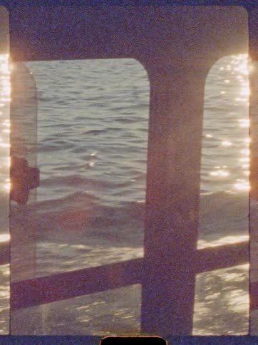The video shows a closeup stylized view of the water from inside or behind a frame possibly a boat window or railingsunlight is strongly reflecting and glistening on the gently moving surface of the water giving the footage a bright vintage or gr