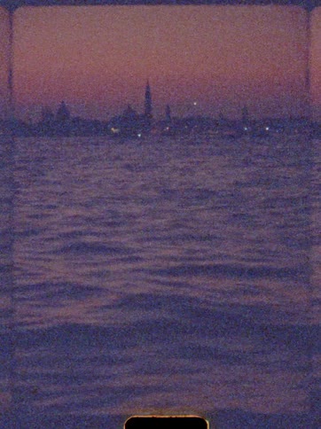 Watercraft with their lights on move across the water near the silhouetted waterfront under a red tinted sky after dusk
