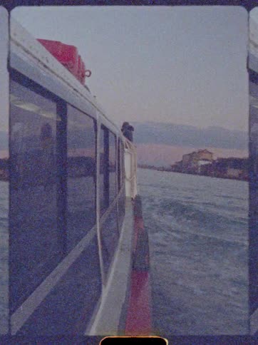 Boat moving along a waterway with an urban shoreline in the background and a person leaning on the side