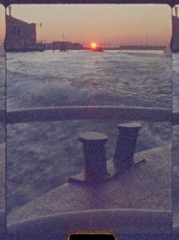 Watercraft moving away from the sun sitting on the horizon while passing the waterfront leaves a wake