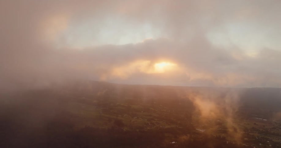 This is an aerial shot showing a landscape shrouded in warm goldenhued mist or low cloudsthe sun is visible as a bright glow breaking through the center of the clouds with beams of light streaming down onto the fields and hills visible below