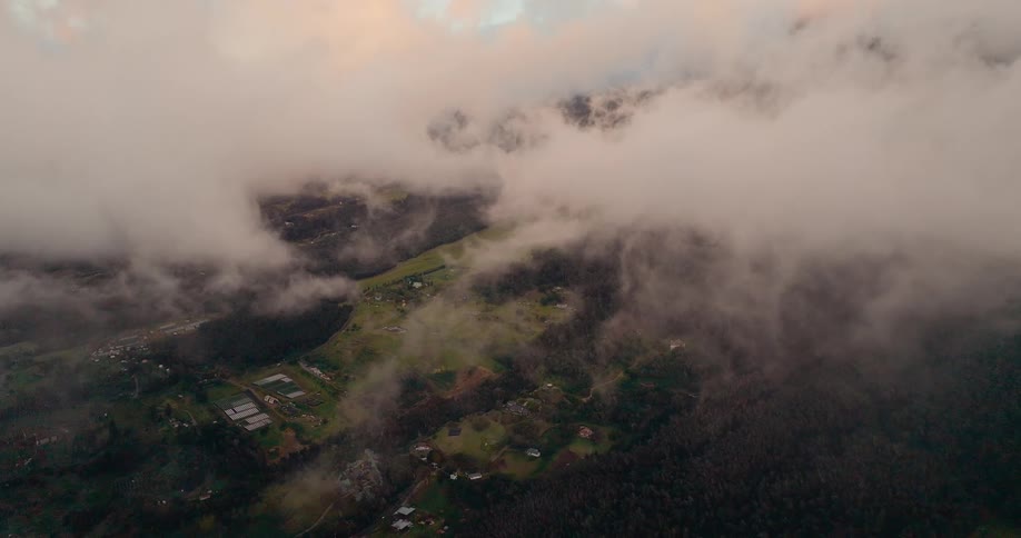 An expansive aerial view of a mountain valley with a mix of forest and scattered homes is mostly covered by heavy slowmoving layers of fog and low clouds