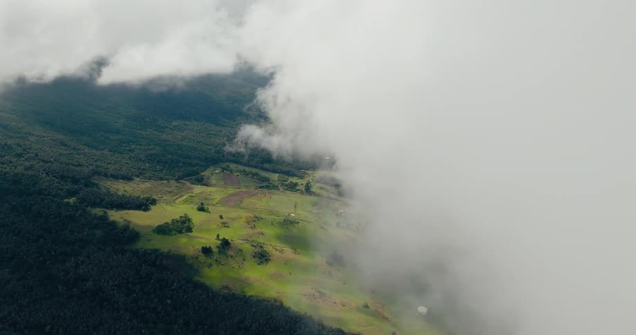This is an aerial view showing thick lowlying white clouds partially obscuring a hillside landscapeas the clouds lift and clear a vibrant green cultivated slope with scattered buildings and dark forests below is revealed