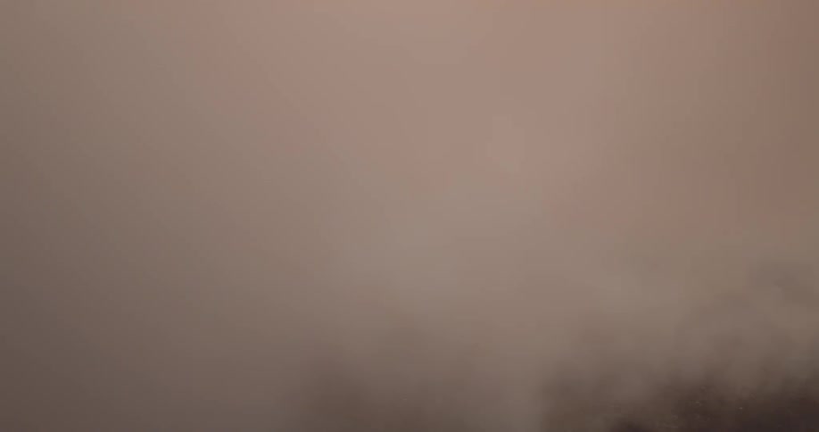 A mistfilled aerial view shows soft clouds drifting across a muted landscape below with warm sunlight breaking through in pale golden and pastel tones that create a calm ethereal atmosphere
