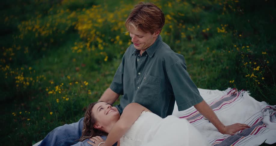 A woman in a white dress is gently lying on a mans lap both relaxing on a picnic blanket in a grassy field dotted with yellow flowers sharing a tender moment