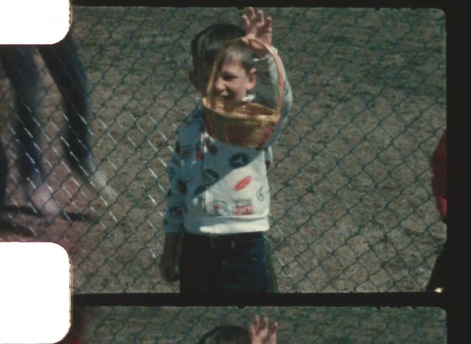 The user wants a description of the video content limited to two linesa young boy dressed in a patterned shirt stands behind a chainlink fence smiling and holding a small woven basket up in the air another person wearing red is partially visib