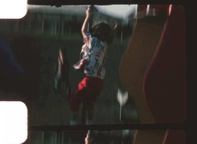 The video shows a child with long hair wearing a patterned top and red pants holding onto a rope or pole and swinging down they appear to be on a piece of playground equipment possibly a slide or a zip line structure
