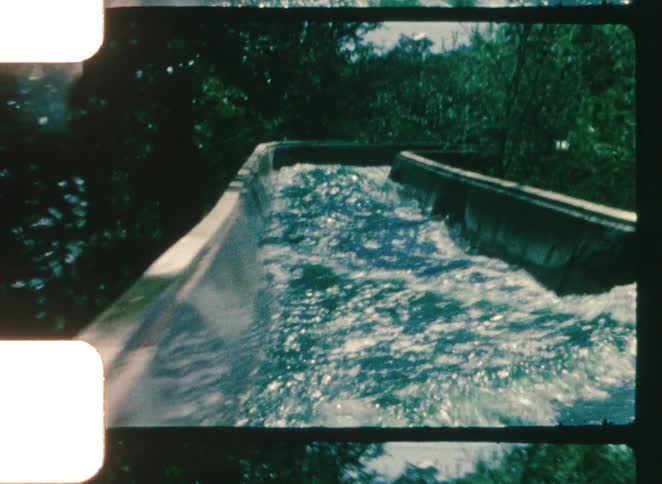 A vintage film clip shows foamy water flowing rapidly through a narrow concrete channel framed by dark green foliage and the distinct film border