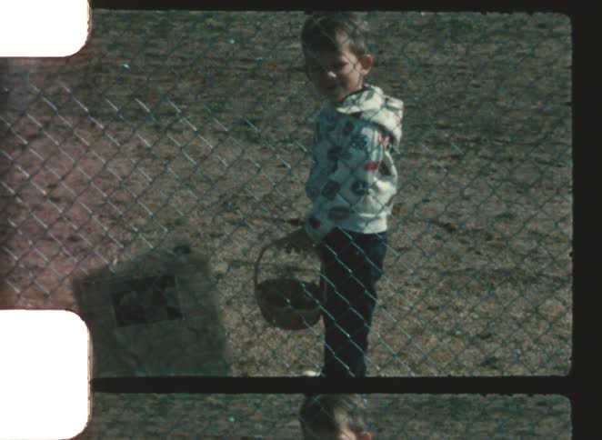 A child wearing a hoodie jacket stands on a grassy field at midday carrying a basket while positioned just beyond a wiremesh fence that runs along the edge of the grassland