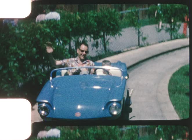 A father enjoying a joyride in an amusementpark car at midday waving as he rides past with his son seated beside him