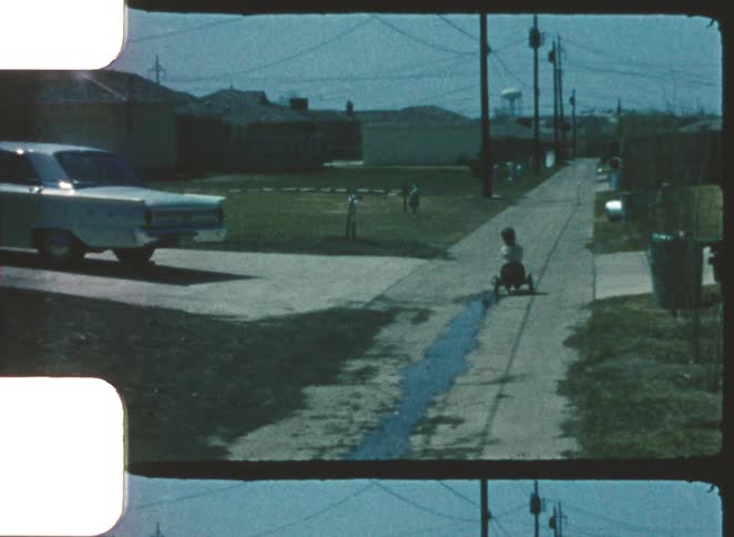 Child rides tricycle in residential alley with vintage car parked and tower in the background