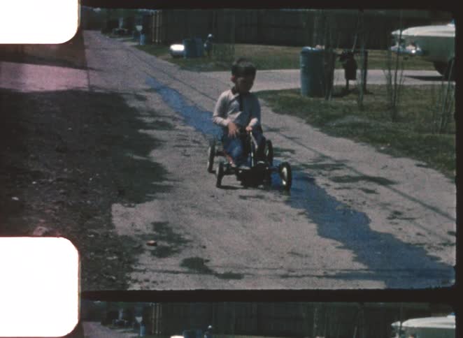 Child riding a pedal cart along a narrow street with puddles of water
