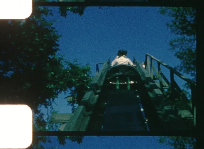 A group of people rides a log flume at an amusement park at midday above the scene an airplane passes across the bright sky