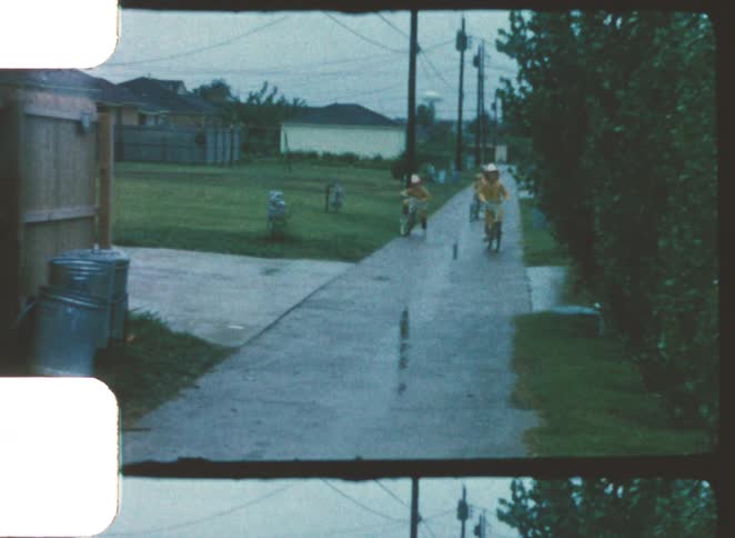 Children riding bicycles on the wet sidewalk in a residential area during light rain