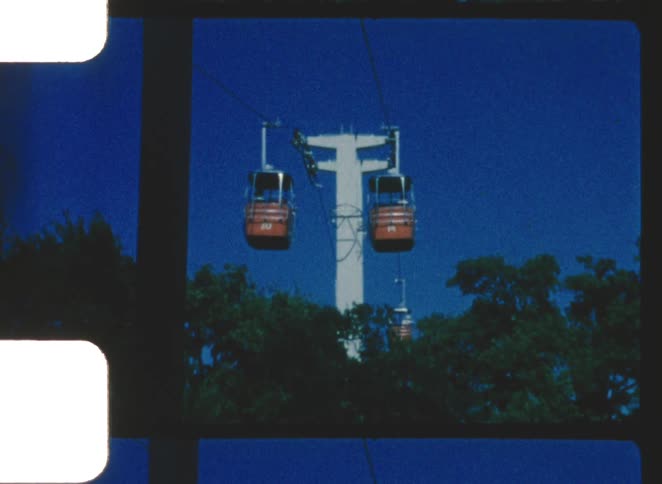 Captured on vintage film with visible sprocket holes bright orange gondola buckets glide along suspended cables supported by a white pylon floating above green treetops against a deep blue sky