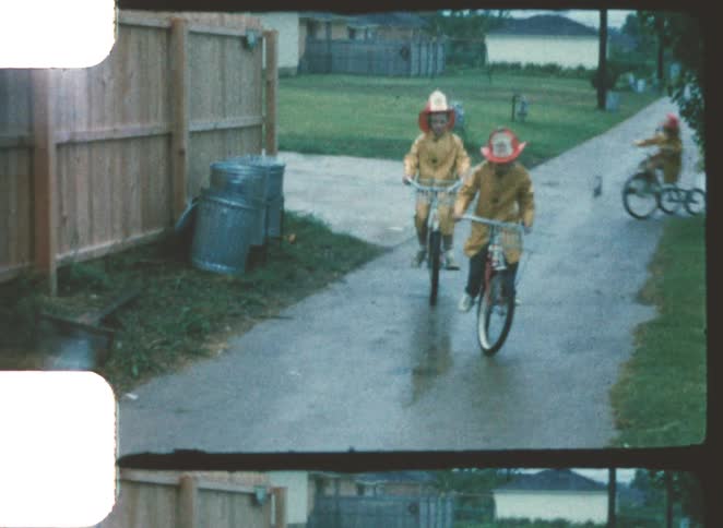 Three young children in yellow raincoats and toy fireman hats happily riding their bicycles down a wet suburban sidewalk on an overcast day