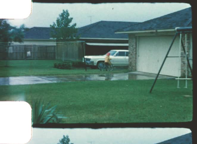 Three children in bright hats ride small bicycles down a wet residential driveway next to a parked white car and a garage