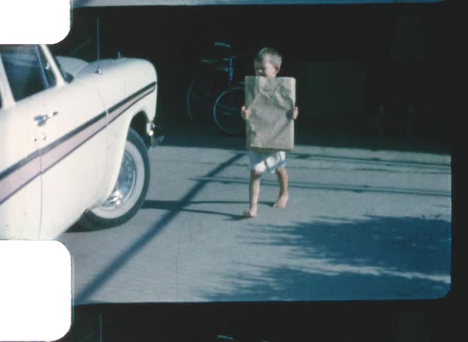 Child walking barefoot along the street carrying a paper bag beside a parked car