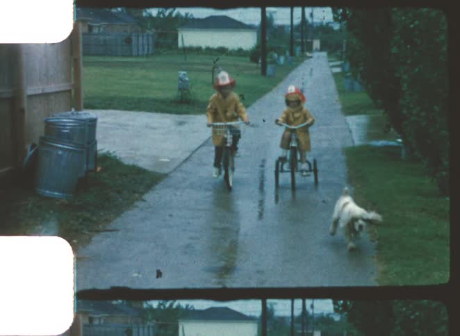 Two young children in yellow raincoats and toy firefighter hats riding a bike and a tricycle with a dog on their side down a wet suburban sidewalk