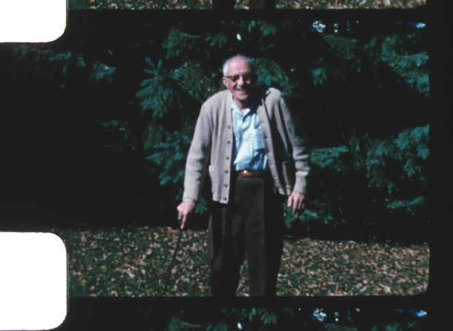 An elderly man wearing a brown jacket over a blue shirt black trousers and glasses walks excitedly across a sunny grassland using a cane as he moves with lively energy