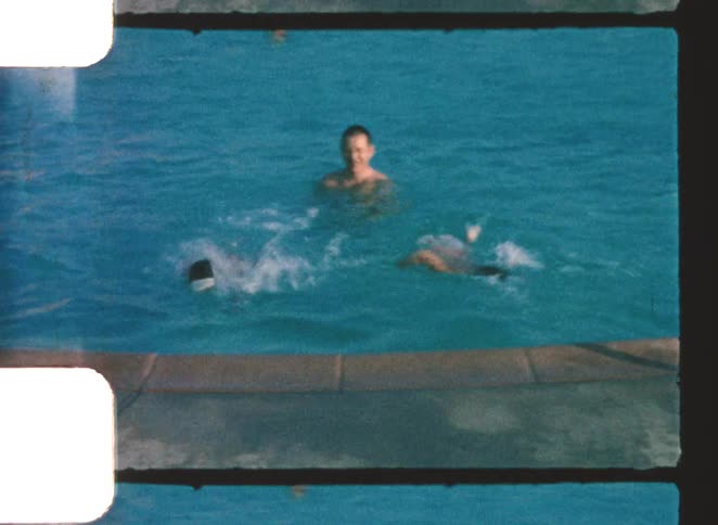 A young boy in red trunks jumps from the edge of a vintagestyle outdoor pool into the water where a man and other children are swimming surfacing to play together in the bright sunny water