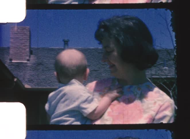 A mother happily holding her bald child standing outdoors on her front lawn at midday