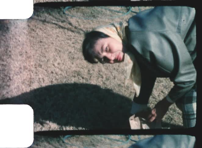 A young woman playfully scooping soil from the ground into a paper bag with one hand wearing a scarf over her head on a sunny day