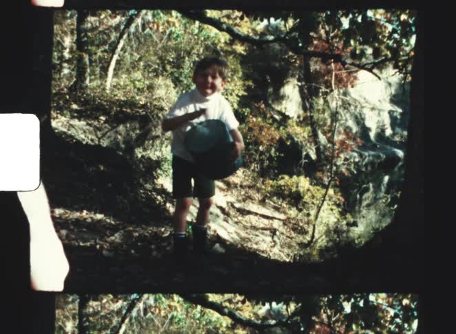 A young child wearing a light shirt and dark shorts stands on a rocky forest path beating on a drum with trees foliage and a rock face surrounding the scene