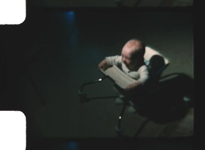 A happy baby in a white sleeper sits in a metal framed infant walker on a dark carpeted floor seen from above in vintage film footage