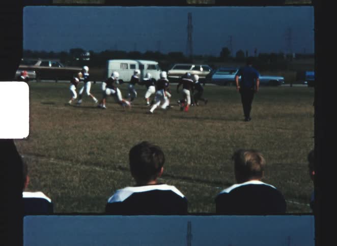 A youth football team in white uniforms with red accents is lined up for a play on a grassy worn field with coaches and several vehicles visible in the far background
