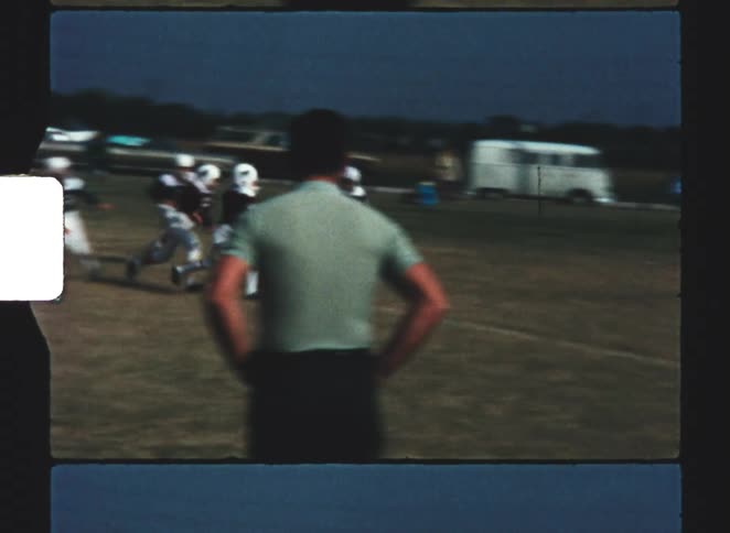Adults watch young football players train in a field near a road lined with vehicles