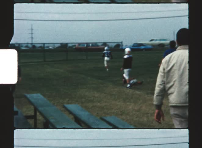 A high angle view shows boys in football uniforms running on a grassy field with a coach in a white jacket watching from the sideline captured in a vintage 8mm film look