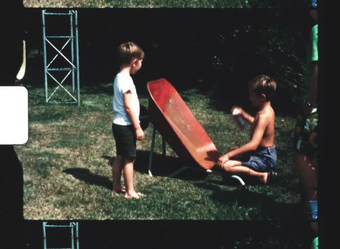 A shirtless kid spray painting a wheelbarrow with other kids watching him outdoors on a sunny grassland