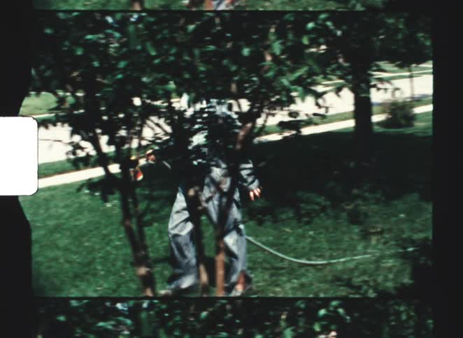 A teenage boy wearing overalls walking on a front lawn holding a spray can and wearing a facemask at midday