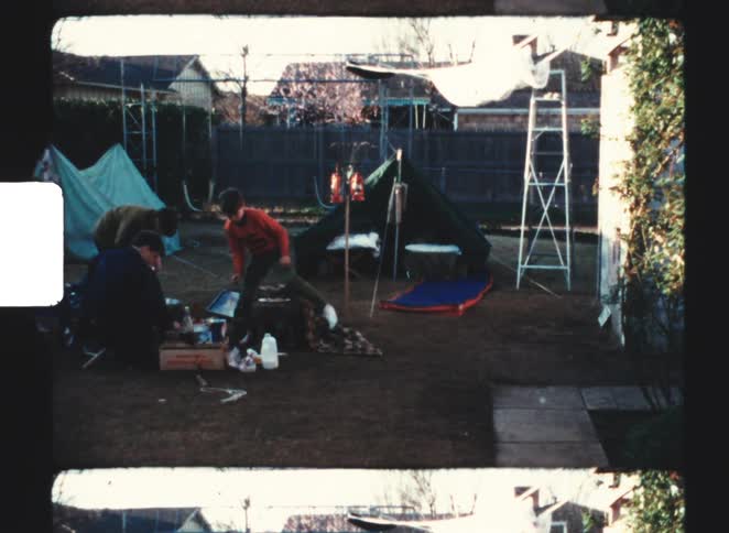 Three people are gathered around camping gear and food in a backyard with two tents and a ladder
