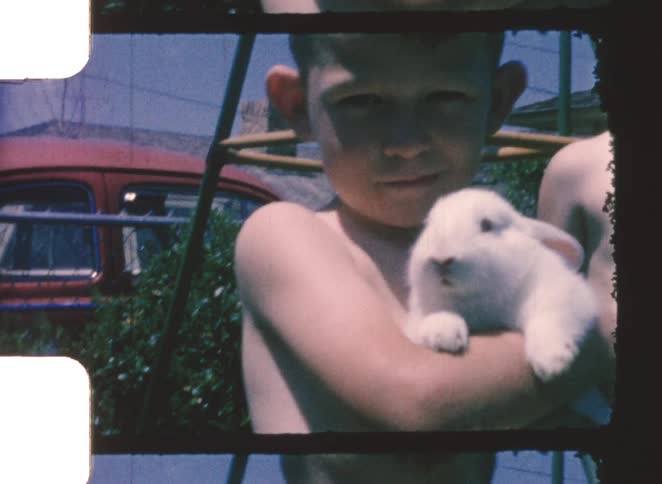 A young boy standing outdoors at midday shirtless and holding a rabbit with another shirtless boy standing beside him