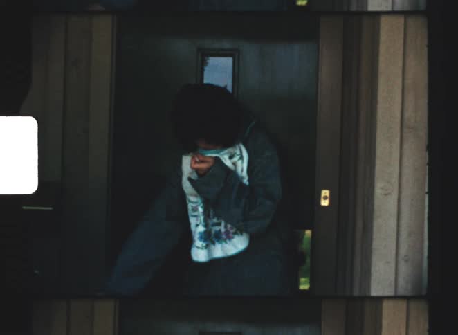 In a candid moment captured on film a young person stands by a wooden door watering a green bush with a garden hose while clutching a white towel around their neck