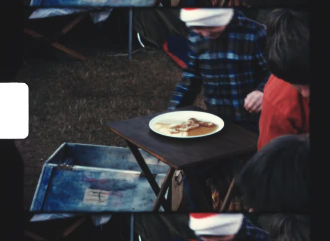 A young boy sitting on a chair during a picnic with friends enjoying his meal while wearing a red beanie hat