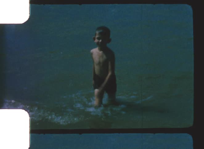A boy is seen shirtless and in dark swim trunks splashing forward through murky water in vintage film footage