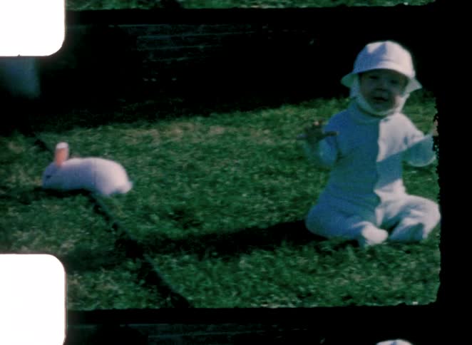 A baby seated on the grass of a backyard with a white rabbit next to him