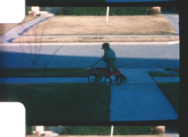 Person walks along sidewalk pulling red wagon in residential neighborhood under soft evening light