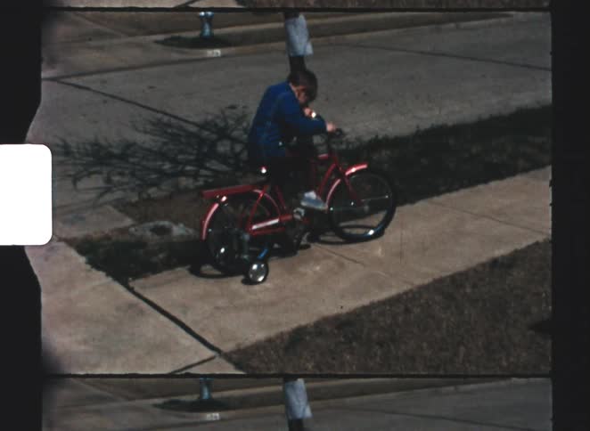 A child being set up by two teenage boys as he rides his red bike with training wheels on the sidewalk on a sunny day