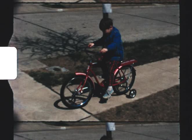 Vintage footage tracks a smiling young boy in a blue jacket pedaling a red bicycle with training wheels down a sunny suburban sidewalk while another child rides alongside him on the grass