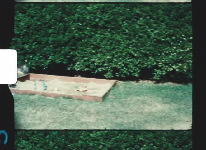 Rectangular sandbox with scattered toys sits on lawn bordered by dense hedge