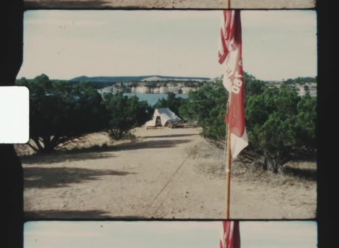 A dirt path leads through low green shrubs toward a white tentlike structure near a body of water in the distance with layered rock formations beyond and a redandwhite flag or banner mounted on a pole in the foreground