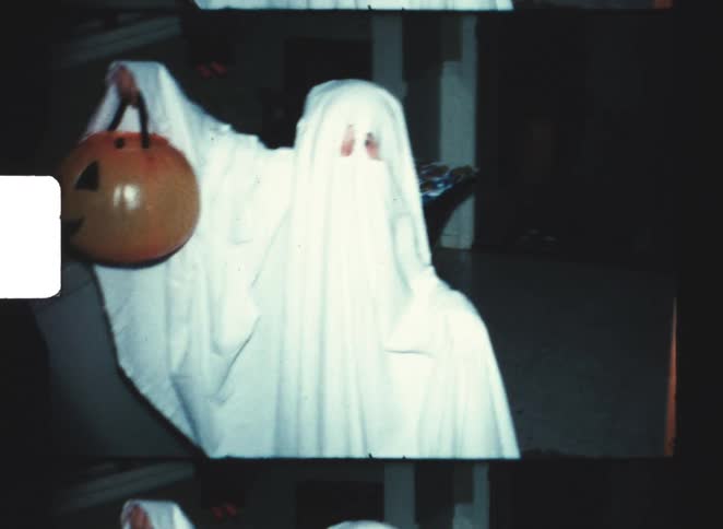 Person dressed as ghost holds pumpkin bucket indoors with staircase in the background